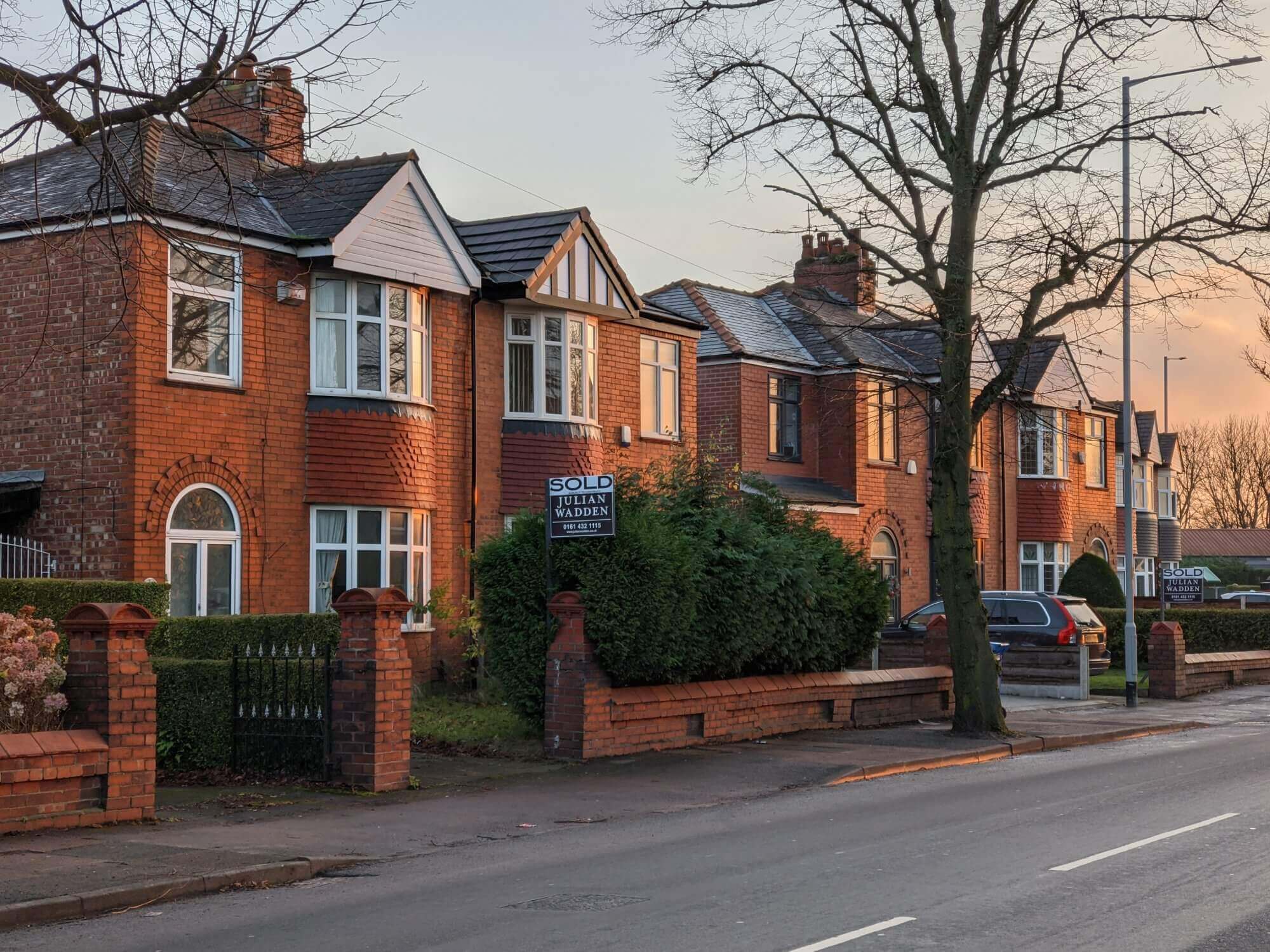 Suburban street with brick houses and a "Sold" sign in front of one property, trees without leaves, and a clear sky. - Home Instead