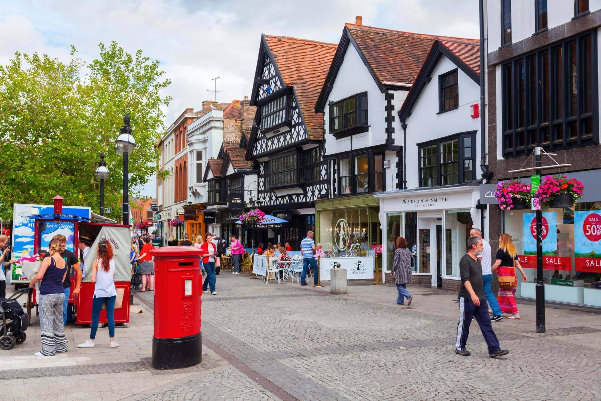 People walking and shopping on a quaint street with half-timbered buildings and a red mailbox in the foreground. - Home Instead