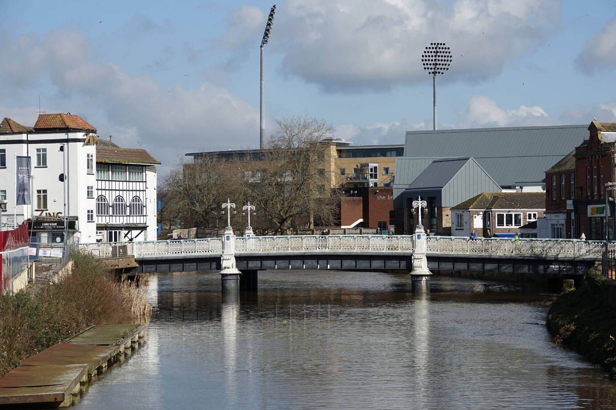 A white bridge spans a calm river with buildings and floodlights in the background under a partly cloudy sky. - Home Instead