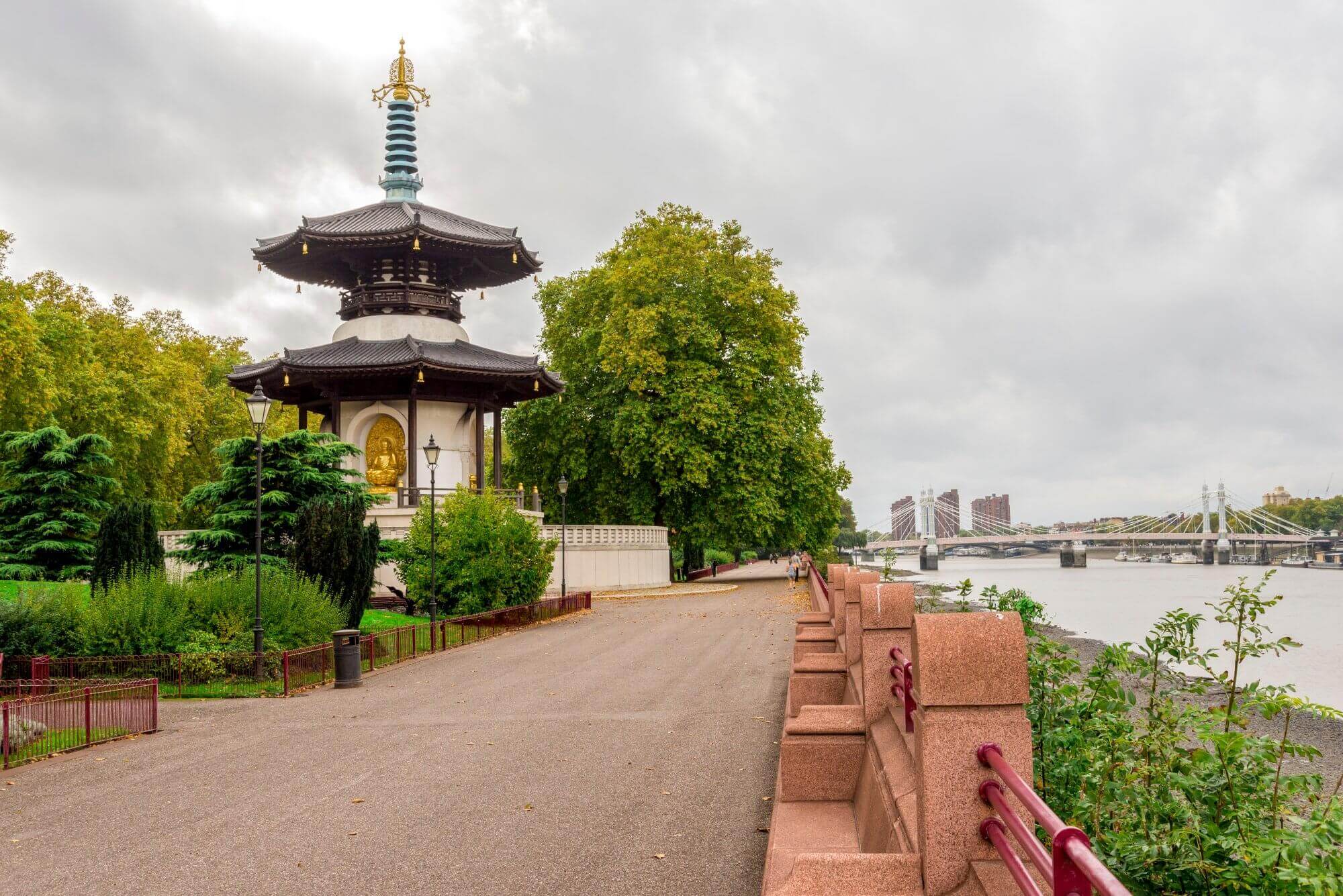 Pagoda near a riverside path with trees around and a bridge visible in the background on a cloudy day. - Home Instead