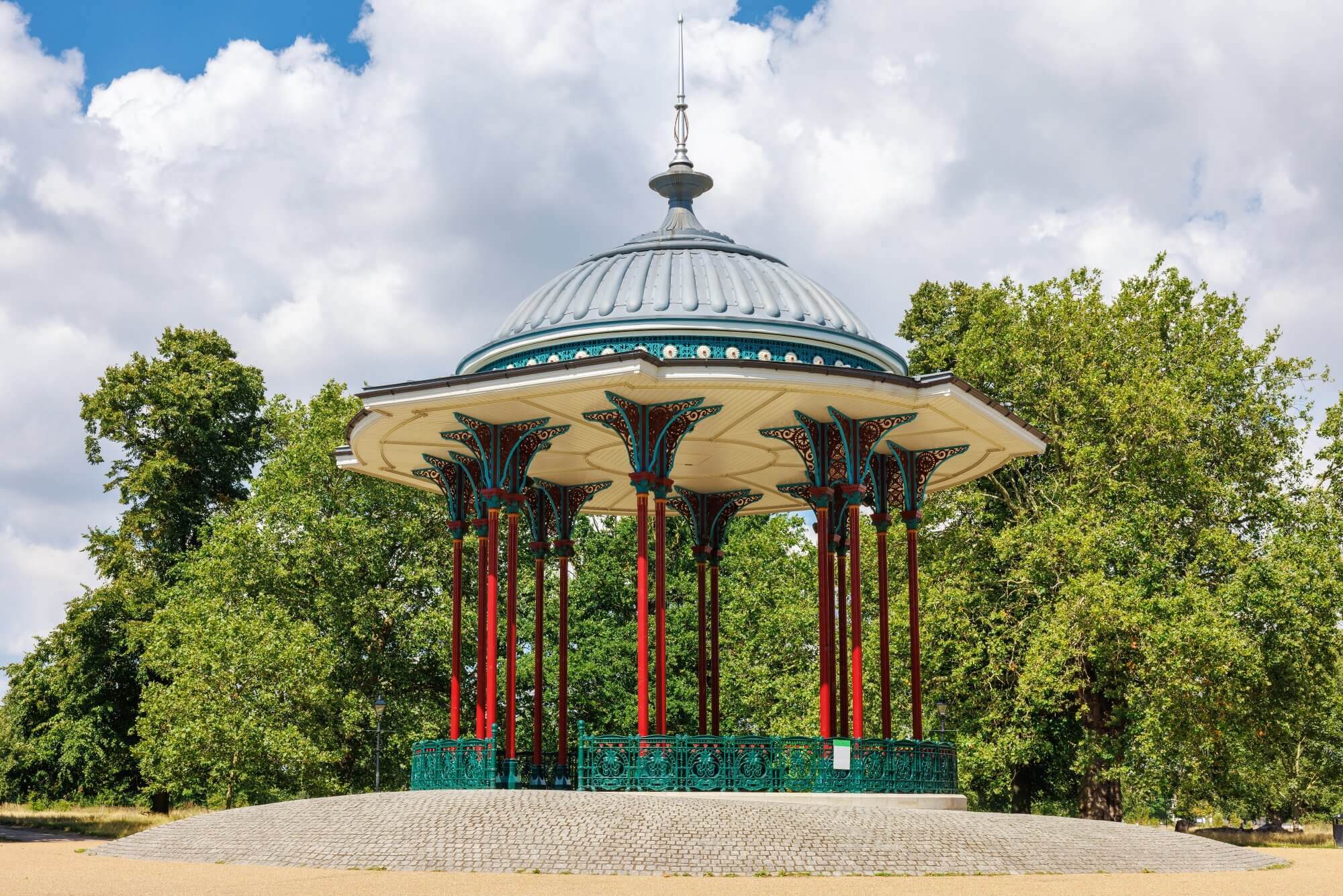 A decorative gazebo with a domed roof and red pillars in a park, surrounded by trees under a cloudy sky. - Home Instead