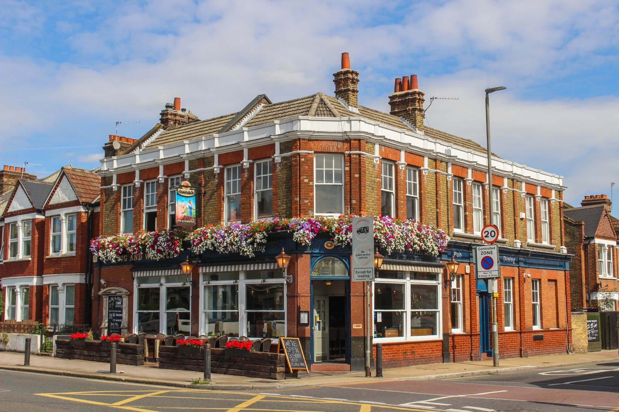 A charming brick pub on a corner street decorated with hanging flowers and a sign indicating a 20 mph speed limit. - Home Instead