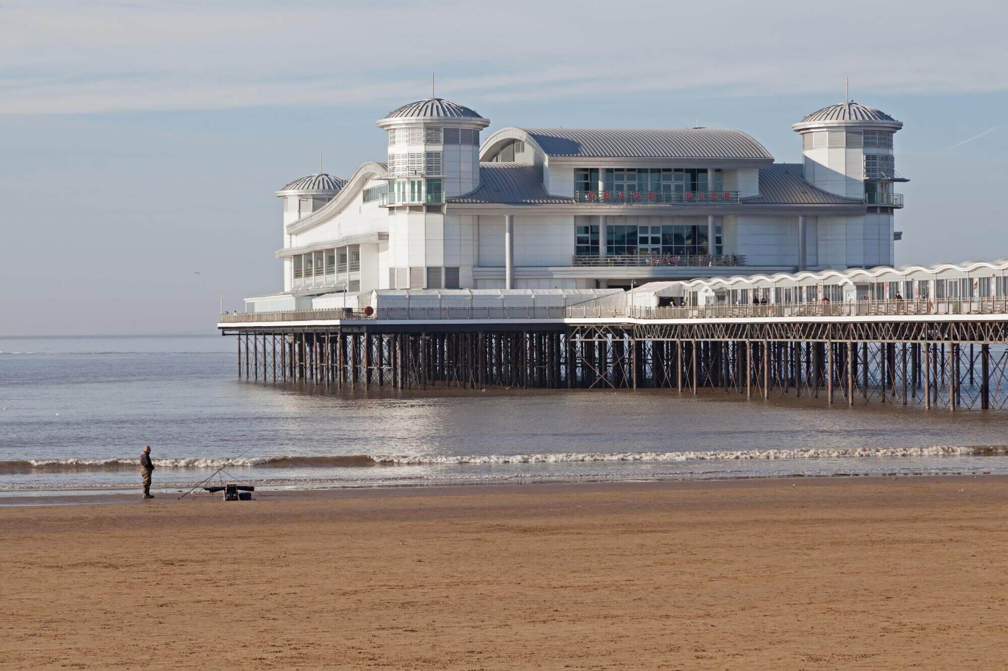 A grand pier with a large white building extends over calm sea waters, while a person and a dog walk on the sandy beach. - Home Instead