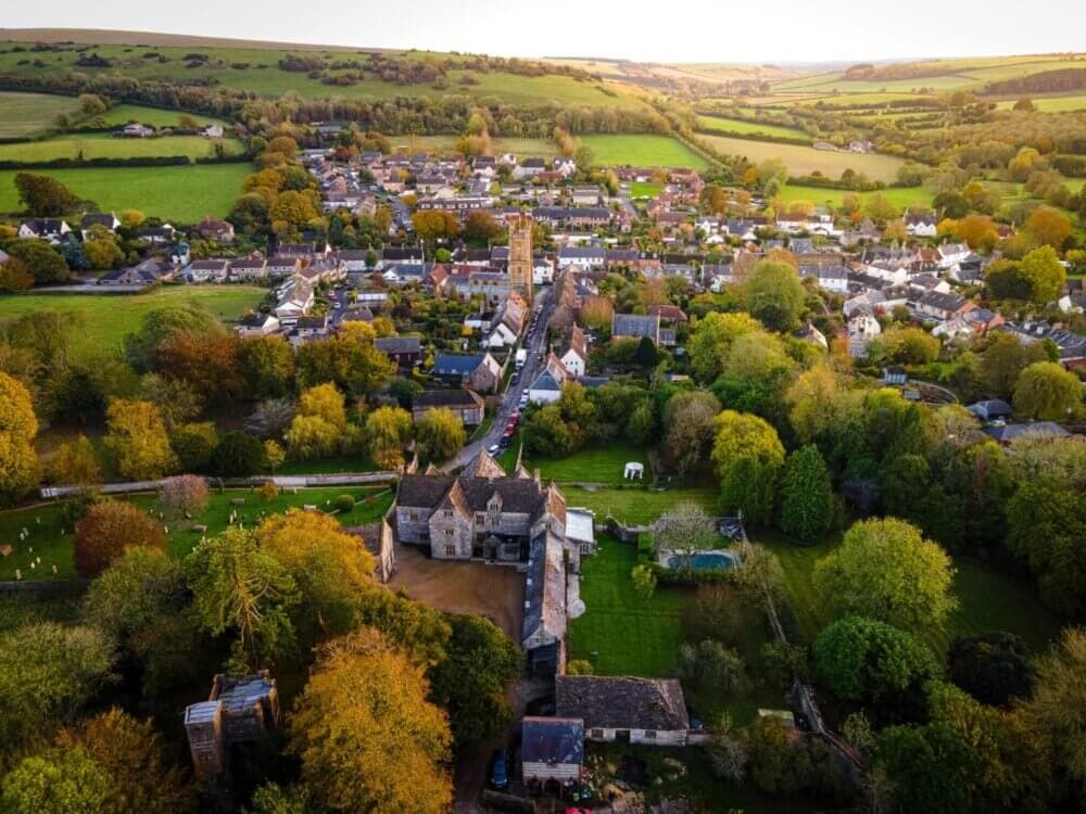 Aerial view of a quaint village surrounded by lush green fields and rolling hills, with a mix of buildings and tree cover. - Home Instead