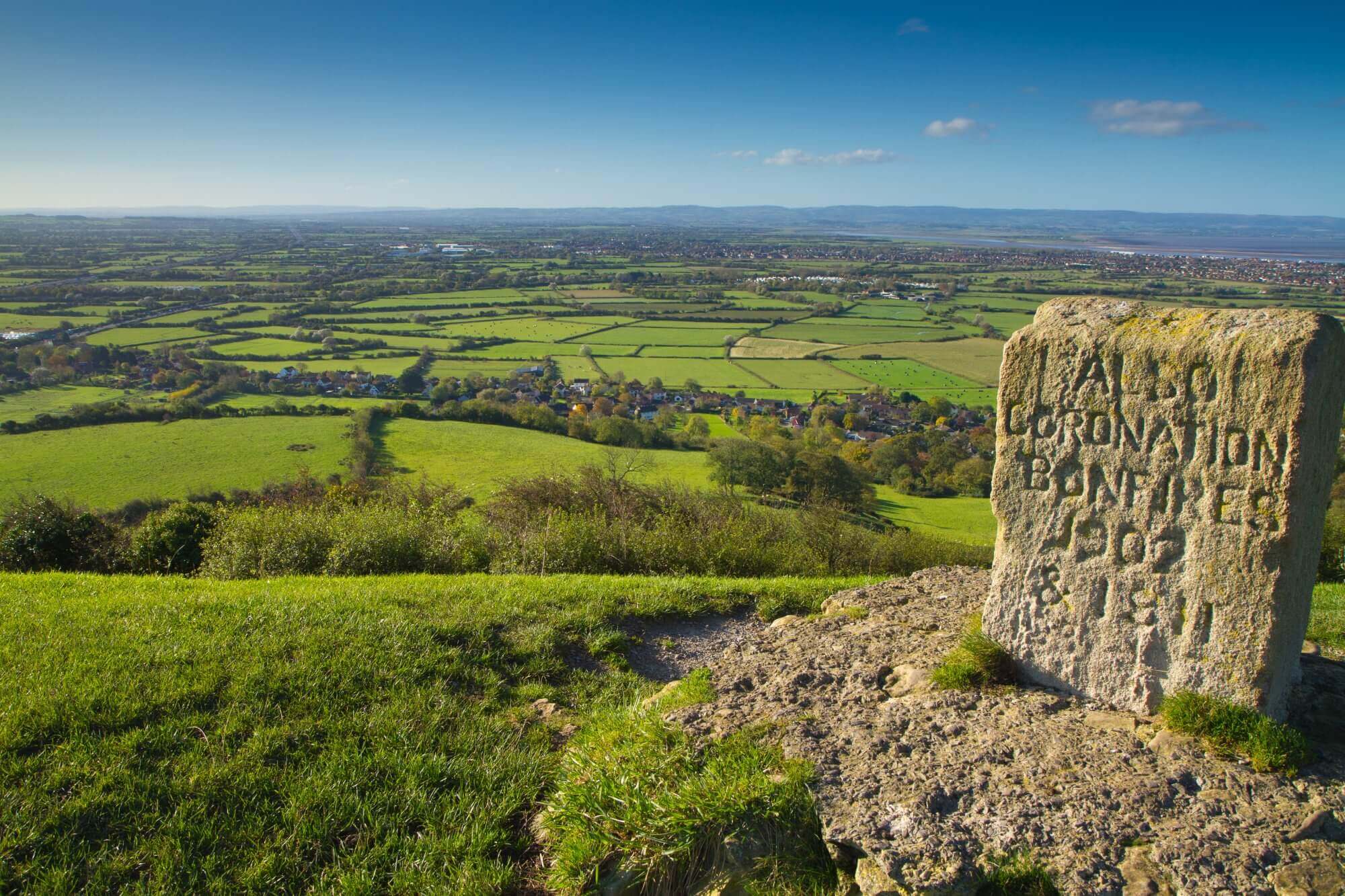 Stone marker with inscription on grassy hill overlooking a vast landscape of fields, houses, and distant hills under a blue sky. - Home Instead