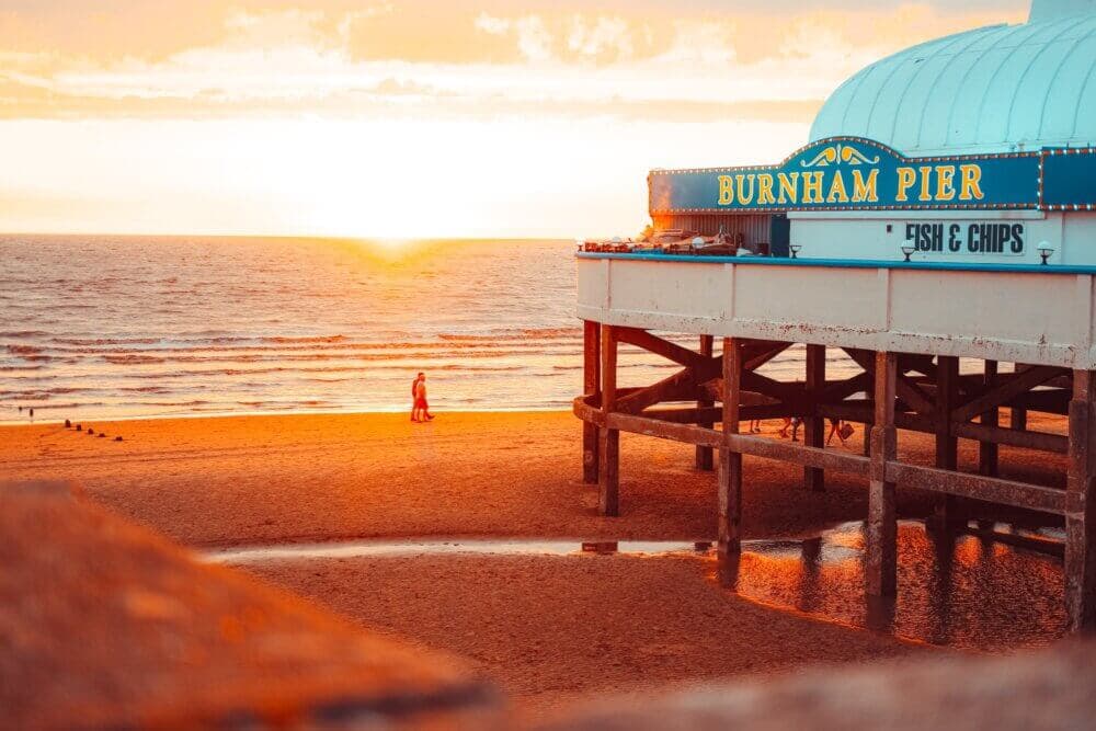 Sunset view of Burnham Pier with a person walking on the beach. The pier sign reads "Burnham Pier" with "Fish & Chips. - Home Instead