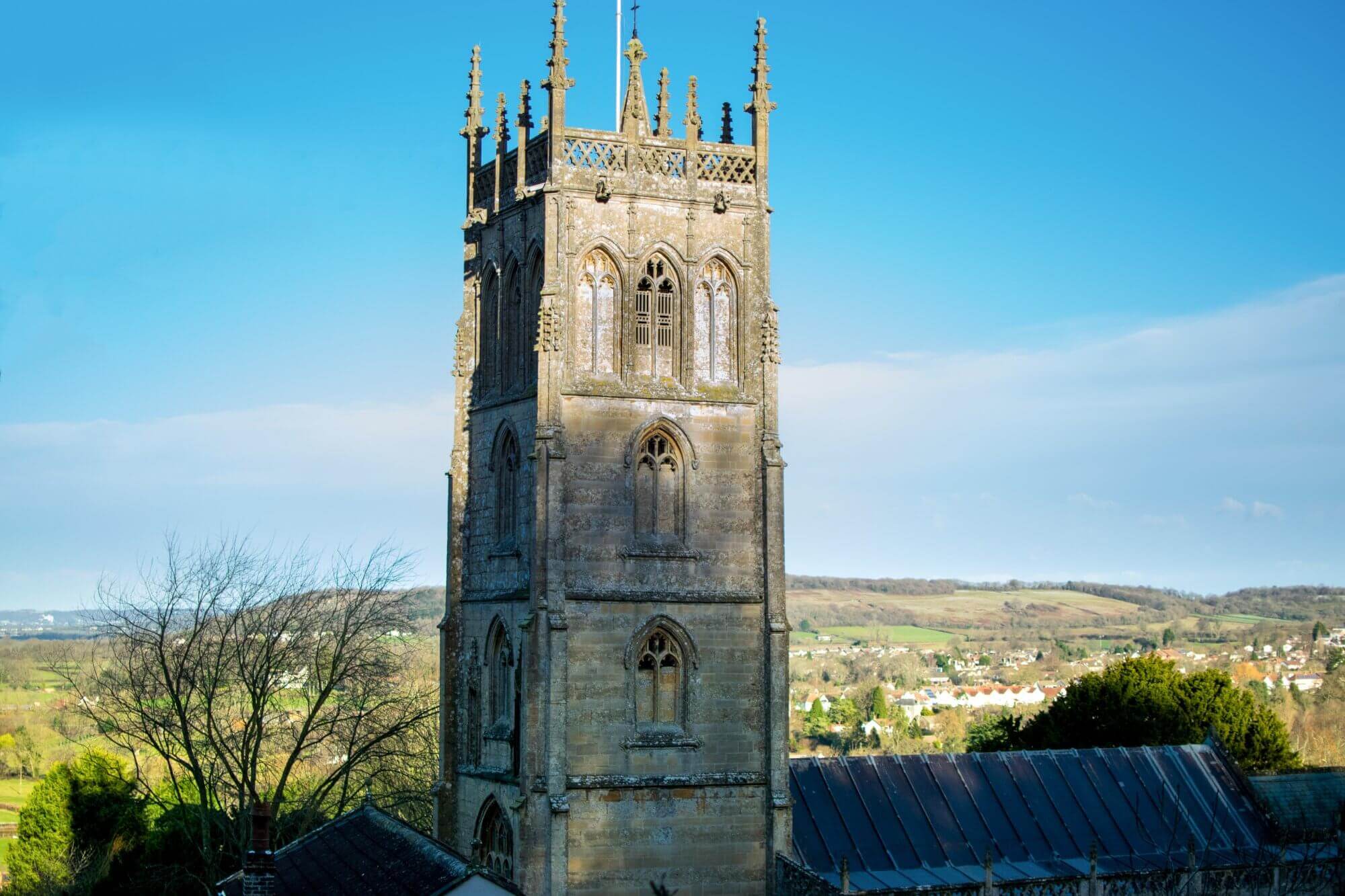 A tall stone church tower stands against a clear blue sky with a countryside landscape in the background. - Home Instead
