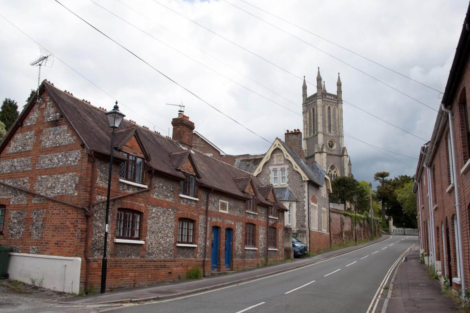 A narrow street lined with brick houses leads to a church with a tall tower under a cloudy sky. - Home Instead