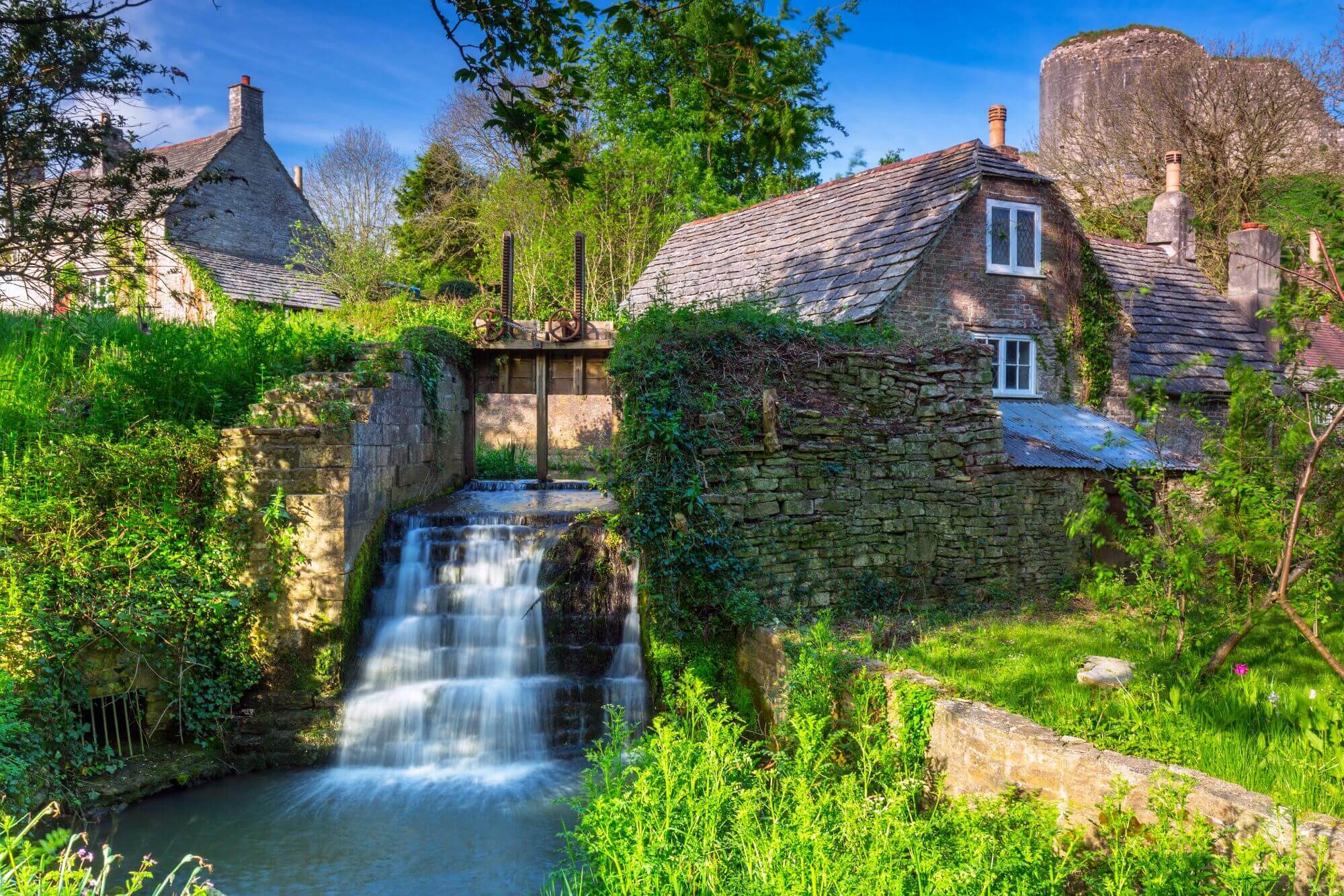 A charming stone cottage beside a small waterfall surrounded by lush greenery under a clear blue sky. - Home Instead