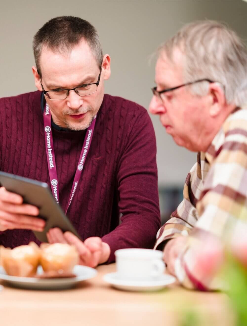 Two older men, one in a red sweater and one in plaid, look at a tablet together at a table with muffins and coffee. - Home Instead
