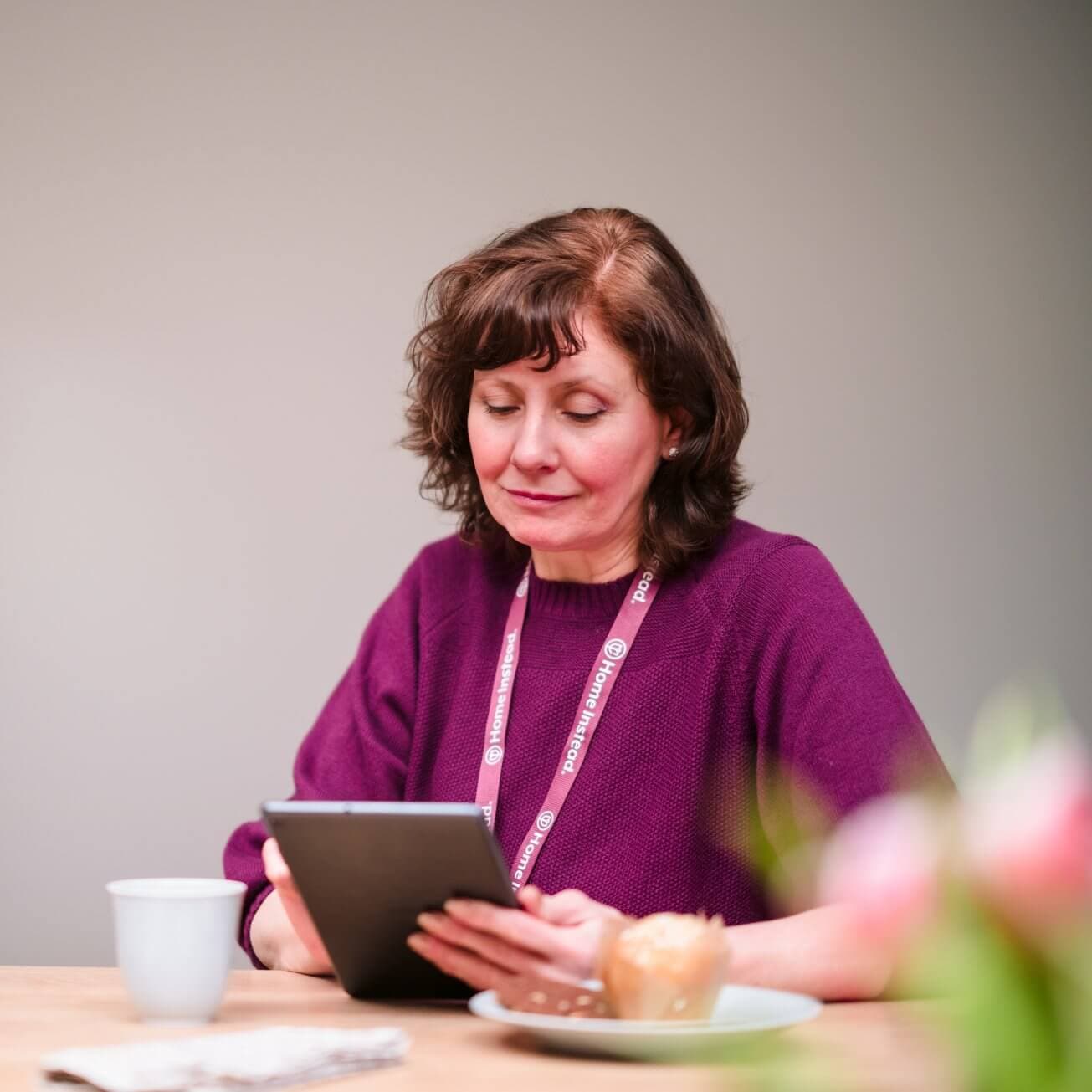 A woman in a purple sweater reads a tablet while sitting at a table with a coffee cup, pastry, and flowers. - Home Instead