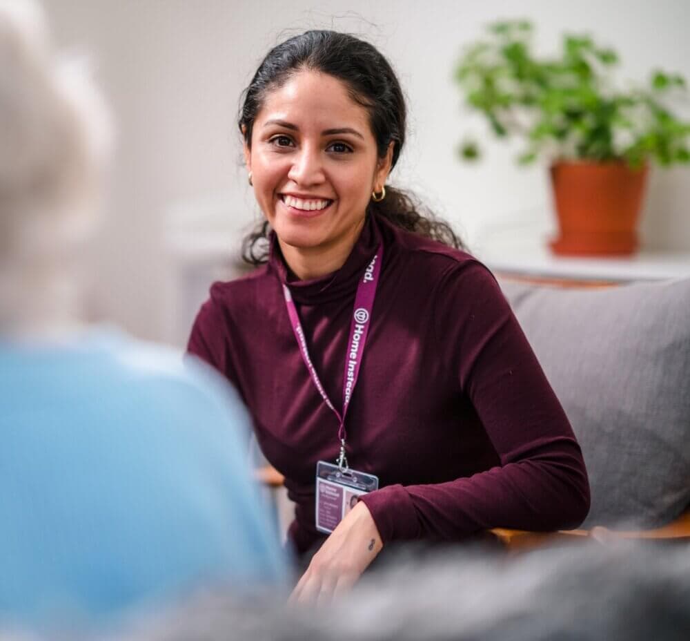 Smiling woman with a name badge and headset, sitting indoors, engaging with an elderly person in the foreground. - Home Instead