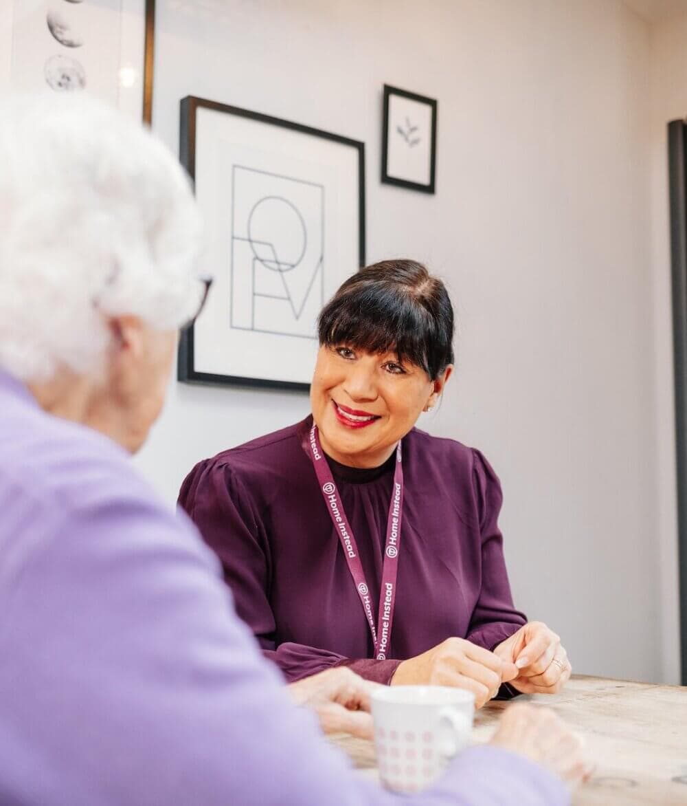 A woman with dark hair talks to an older person, both seated at a table with mugs, in a bright room decorated with artwork. - Home Instead