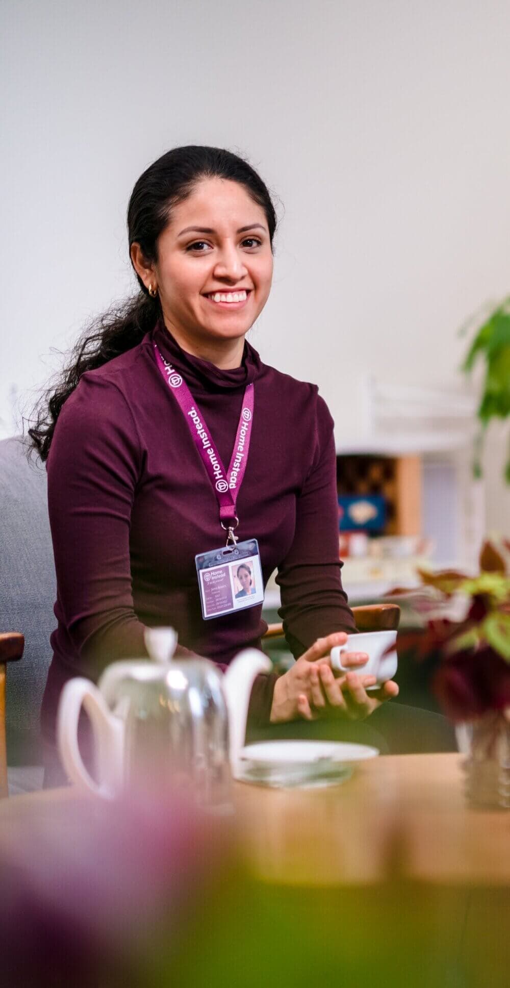 Smiling woman wearing a lanyard and holding a cup, seated at a table with a teapot and plants in the foreground. - Home Instead