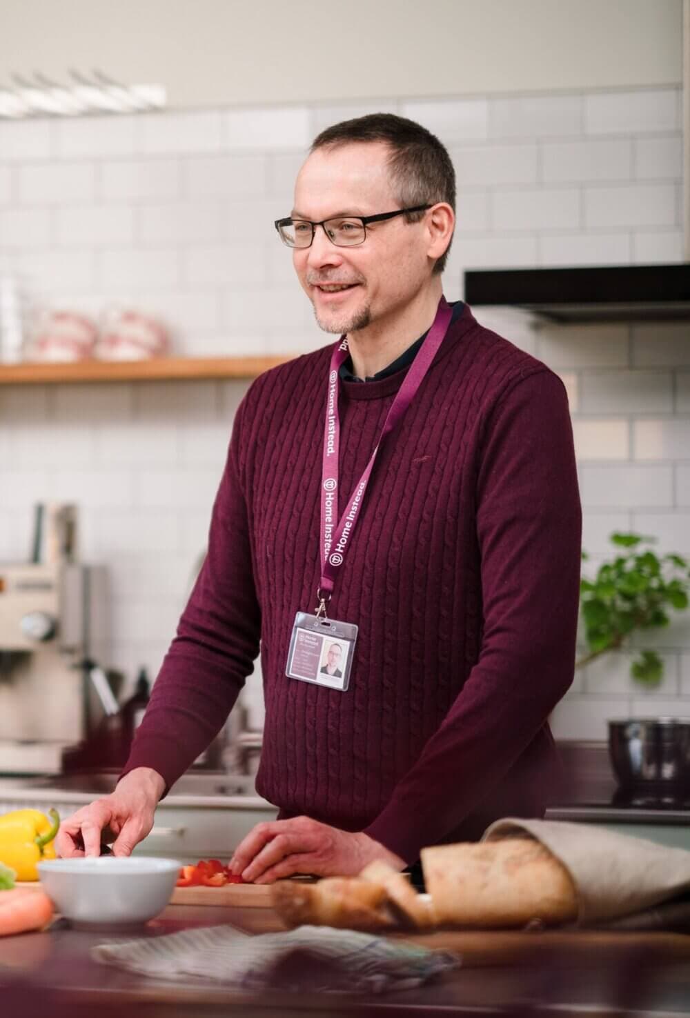 A man wearing glasses and a name badge, slicing tomatoes in a modern kitchen with white subway tiles. - Home Instead