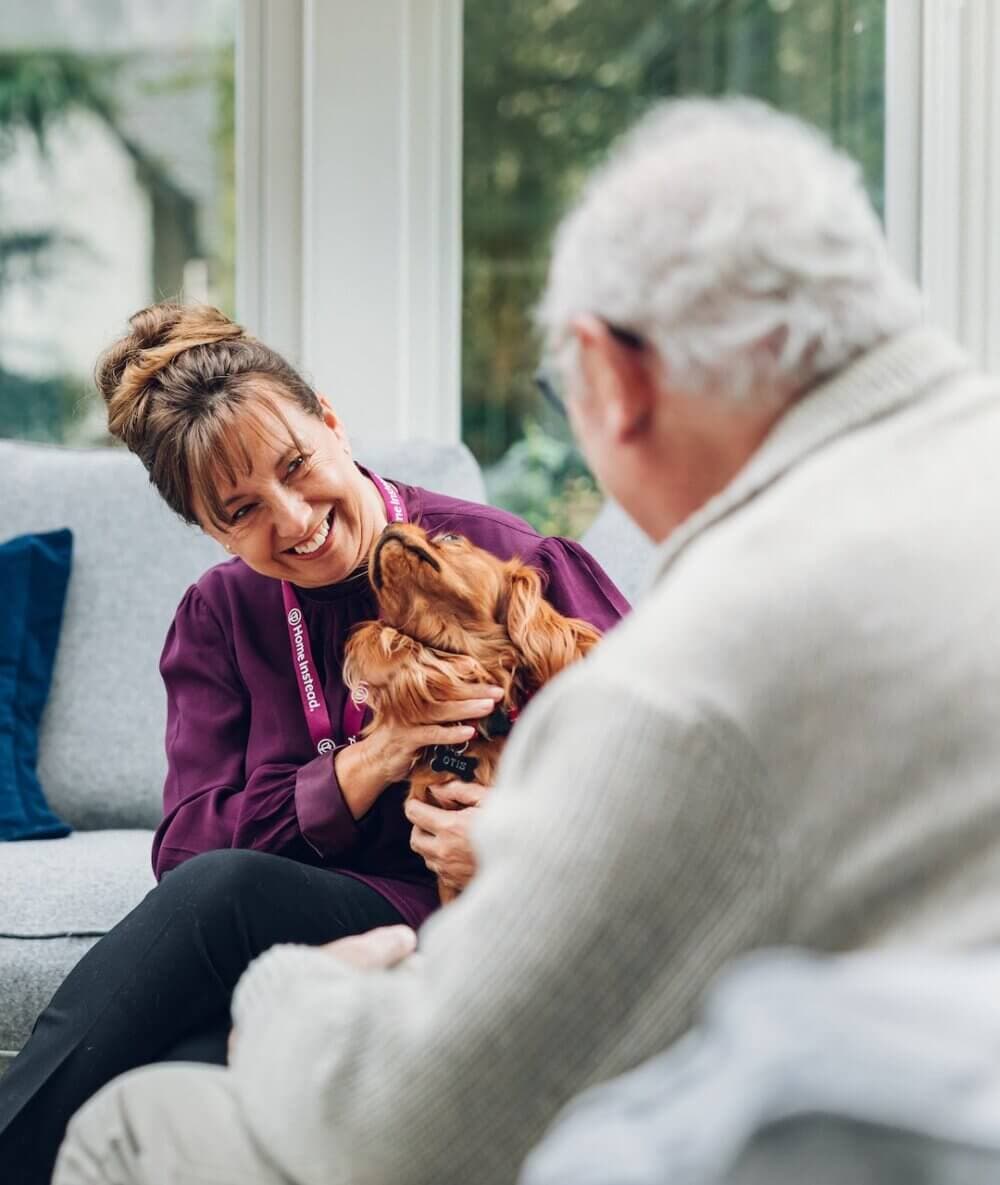 A woman smiling and holding a dog, sitting on a couch with an elderly man facing her. - Home Instead
