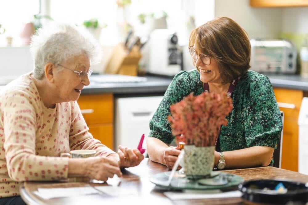 Two women sitting at a wooden table in a kitchen, chatting and looking at papers, with a plant in the foreground. - Home Instead
