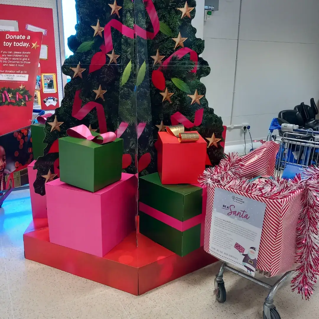 A large Christmas tree display with wrapped gifts, and a shopping cart filled with tinsel and a donation sign. - Home Instead