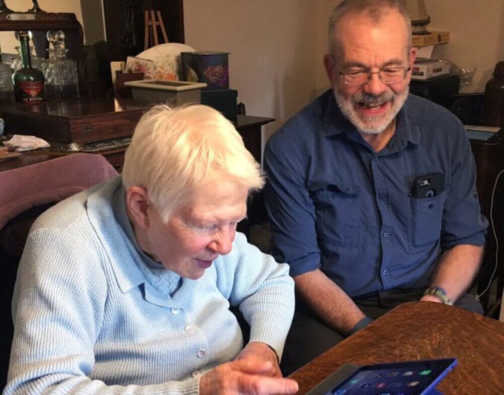 An elderly woman and a man smiling while looking at a tablet together at a wooden table. - Home Instead