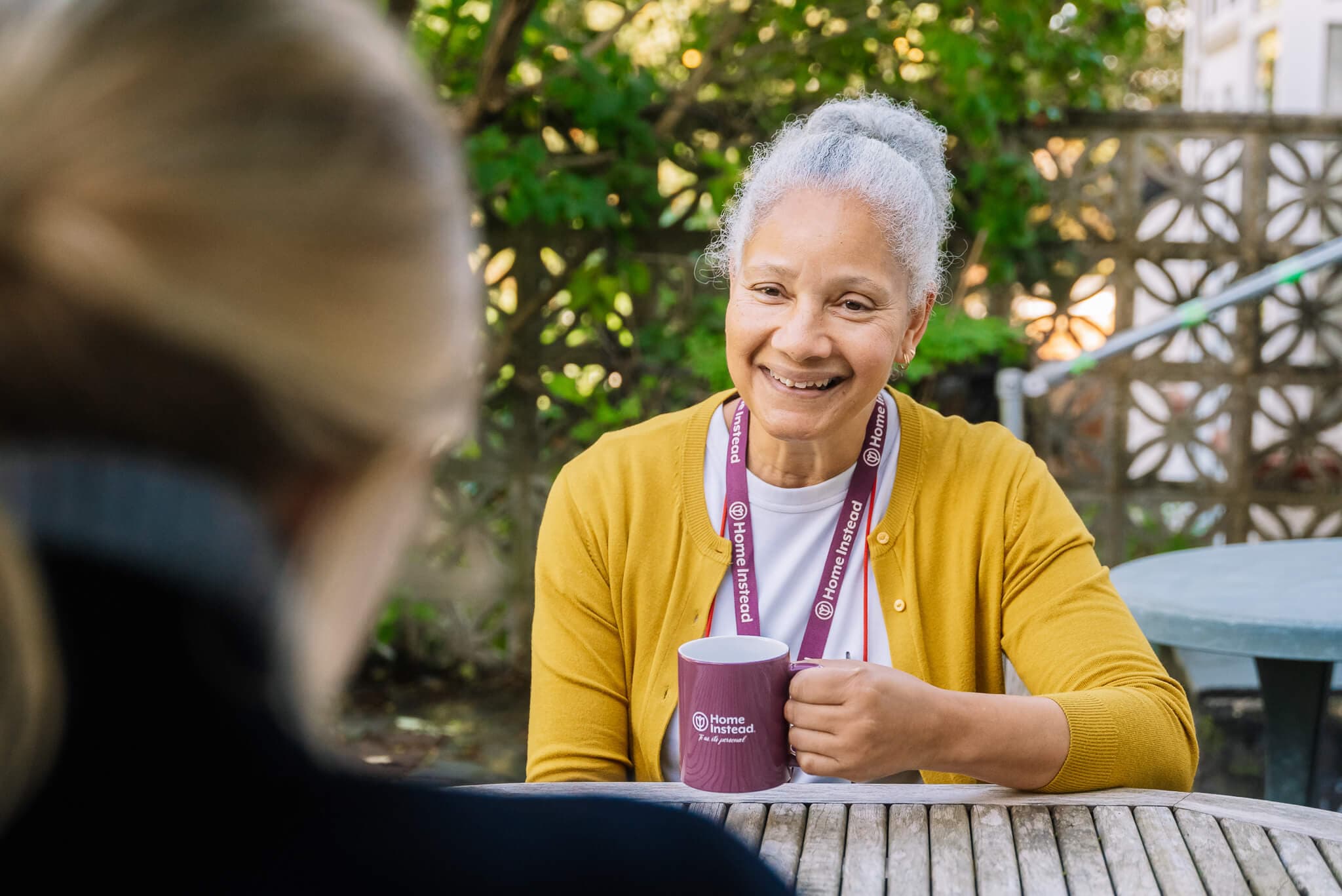 Smiling woman with gray hair holding a mug, sitting at a table outdoors, wearing a yellow cardigan and lanyard. - Home Instead