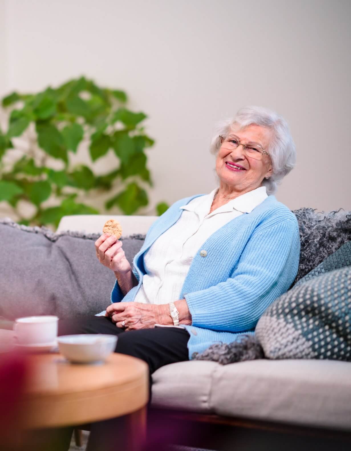 Elderly woman with white hair sitting on a couch, smiling and holding a cookie, with a cup and plants in the background. - Home Instead Southampton