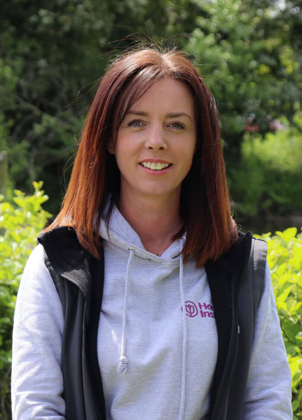 A woman with shoulder-length brown hair wearing a gray hoodie and black vest smiles outdoors with greenery in the background. - Home Instead