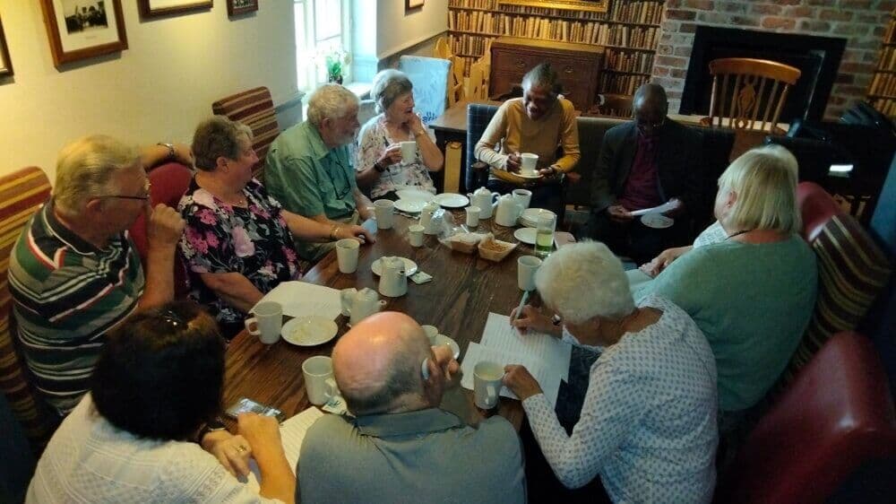 A group of seniors enjoying tea and writing at a cozy wooden table in a room with bookshelves and framed pictures. - Home Instead