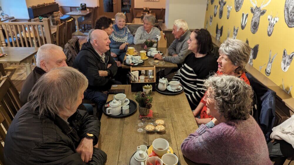 Group of seniors sitting around a wooden table in a cozy café, enjoying tea and pastries, engaged in lively conversation. - Home Instead