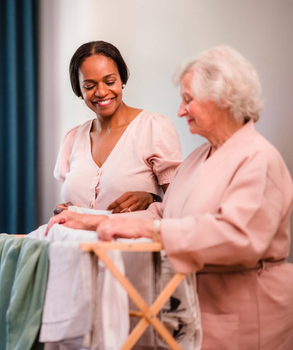 A young woman smiles as she helps an older woman folding laundry on a drying rack. - Home Instead
