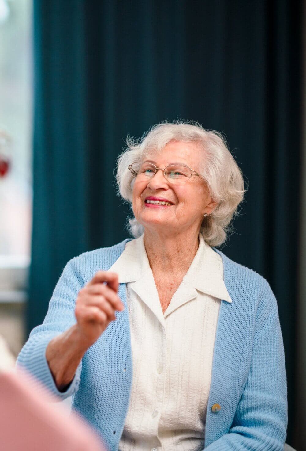 An elderly woman with white hair and glasses, wearing a blue cardigan and white shirt, smiles while pointing at something. - Home Instead