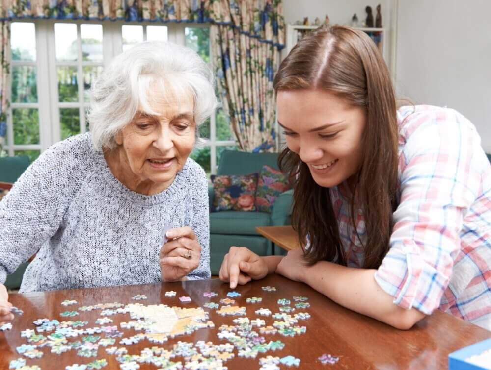 Older woman and young woman smiling while working together on a jigsaw puzzle at a wooden table in a cozy living room. - Home Instead