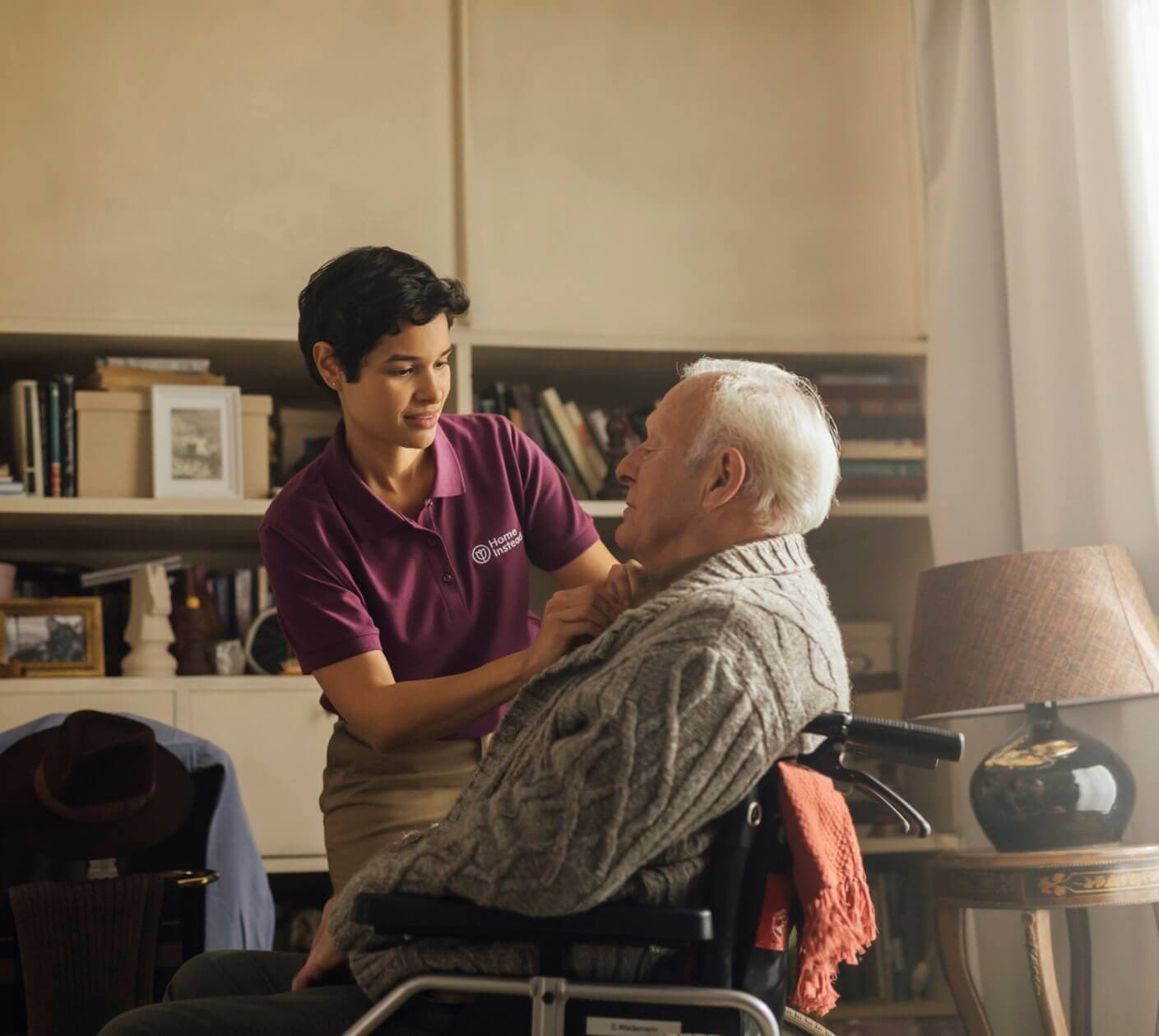 Caregiver in a purple shirt assisting an elderly man in a wheelchair with his sweater in a cozy living room. - Home Instead