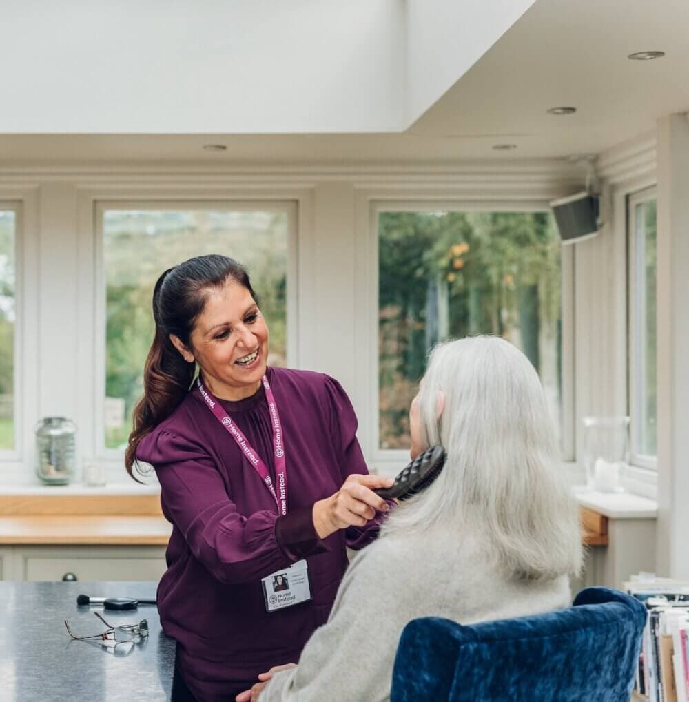 A caregiver gently brushes the hair of a seated elderly woman in a bright room with large windows. - Home Instead