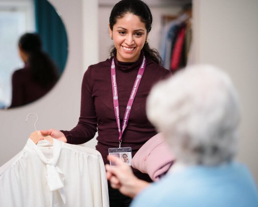 A smiling caregiver shows a white blouse to an elderly woman in a blue top inside a cozy room. - Home Instead