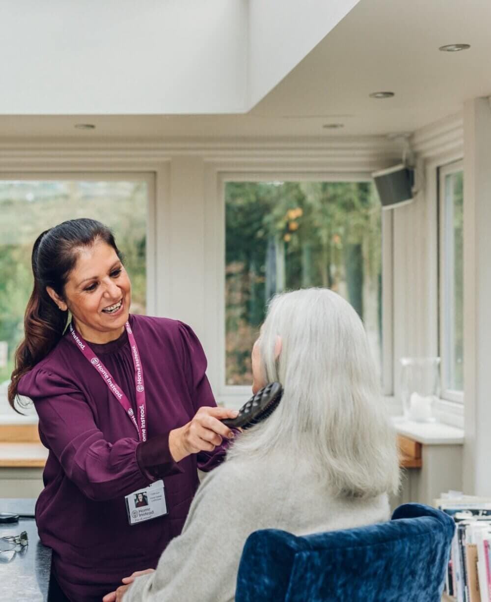 A caregiver is brushing the hair of an elderly woman in a sunlit room with large windows. - Home Instead