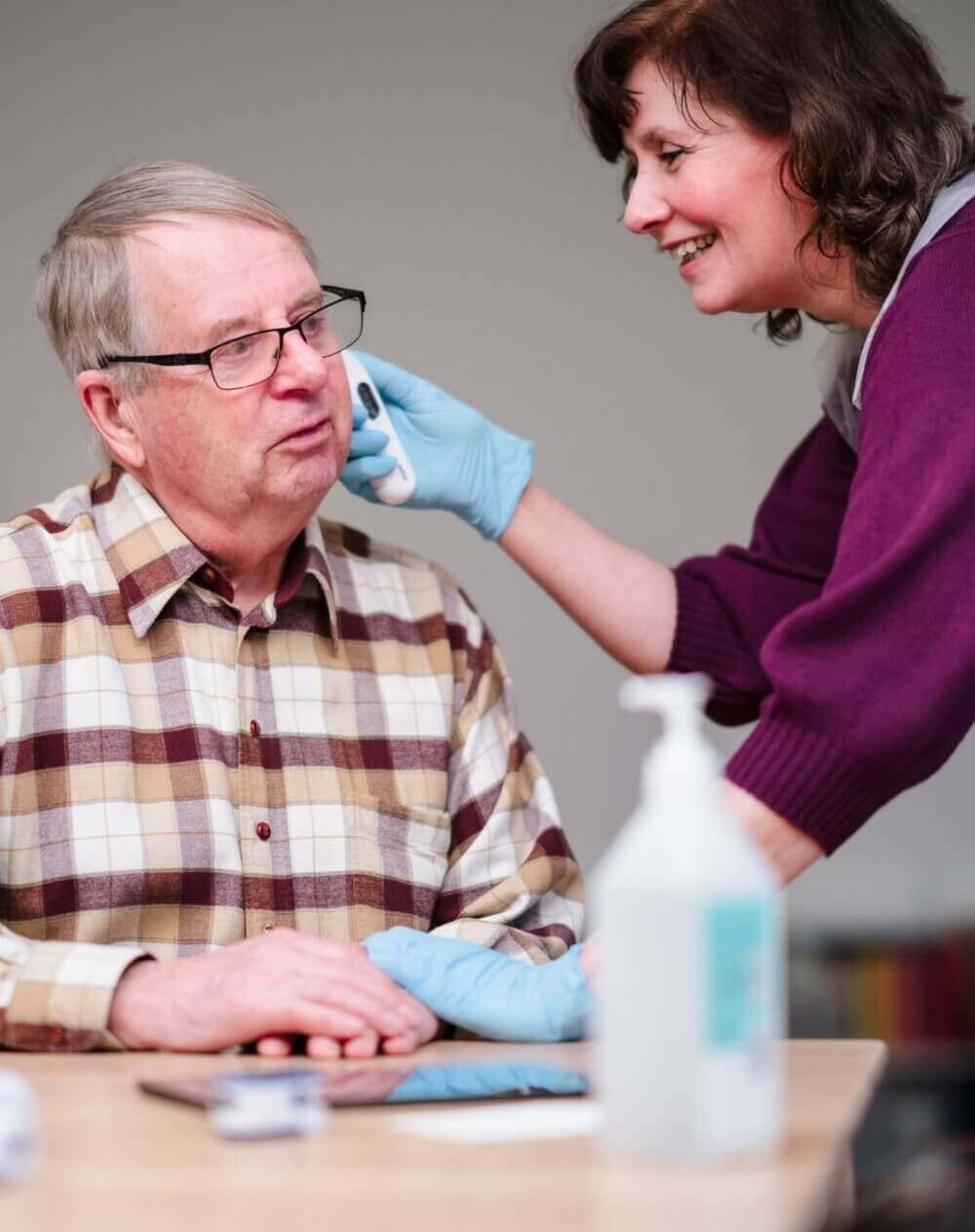A caregiver, smiling, assists an elderly man by cleaning his ear at a table with medical supplies. - Home Instead