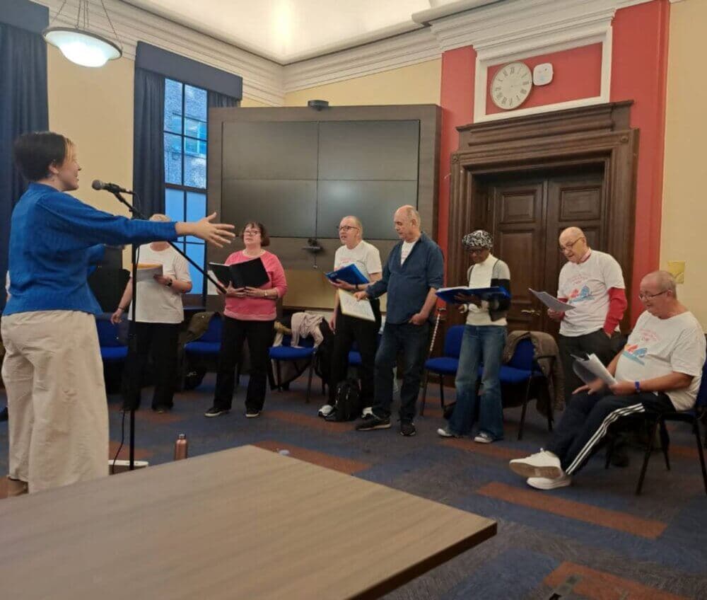 A group of people stands and sits around a central figure directing them, likely rehearsing or performing in a room. - Home Instead