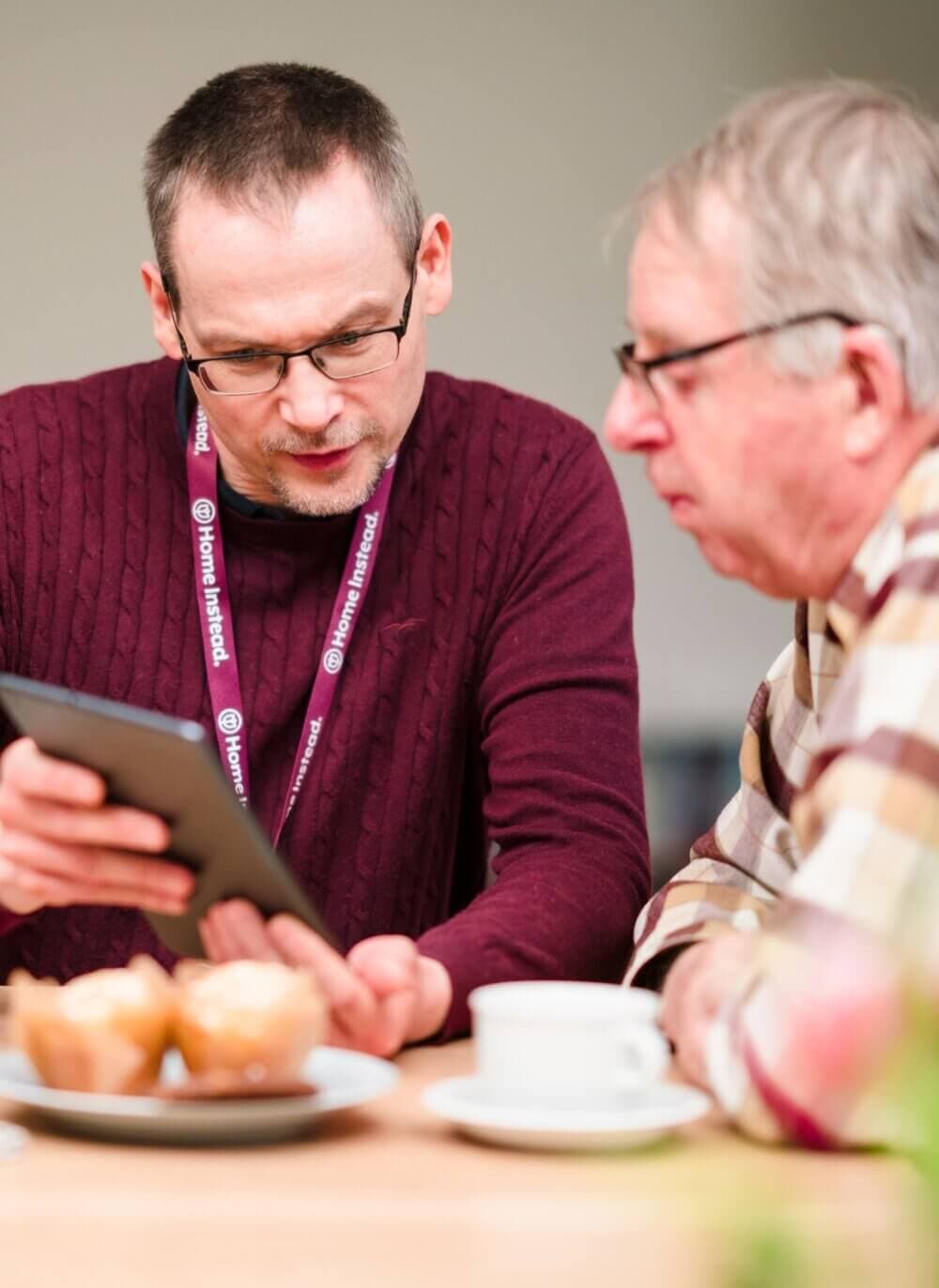 Two men sitting at a table, looking at a tablet. One is wearing a lanyard, and there's a mug and desserts on the table. - Home Instead