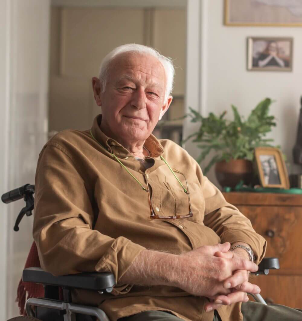 Elderly man sitting in a wheelchair, wearing a brown shirt and glasses, smiling softly with hands clasped together. - Home Instead