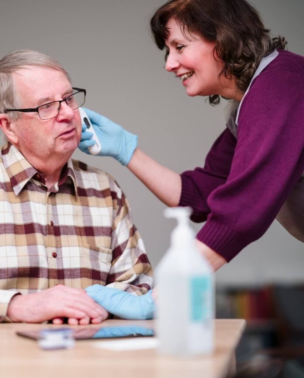 A caregiver in gloves uses an ear thermometer to check an elderly man's temperature at a table. - Home Instead