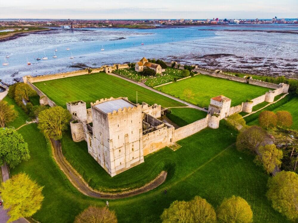 Aerial view of a historic seaside castle with large green lawns, surrounded by trees and water, boats in the distance. - Home Instead