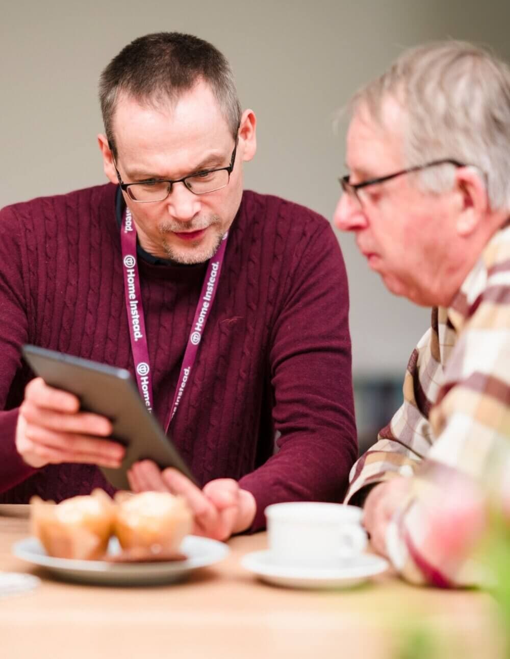 Two men sitting at a table with muffins and a cup of coffee, looking at a tablet together. - Home Instead