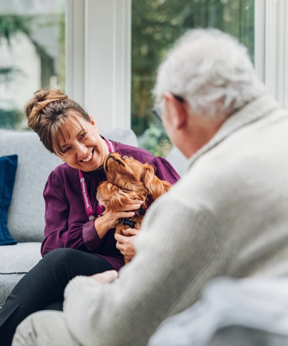 A woman and an elderly man smile as they pet a dog together in a cozy living room. - Home Instead