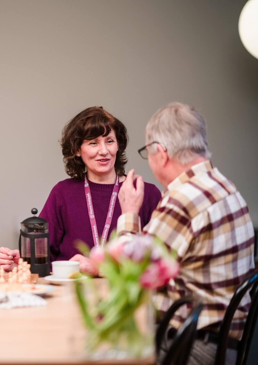 Two people conversing at a table with coffee, a chessboard, and flowers. The woman wears a lanyard and smiles. - Home Instead