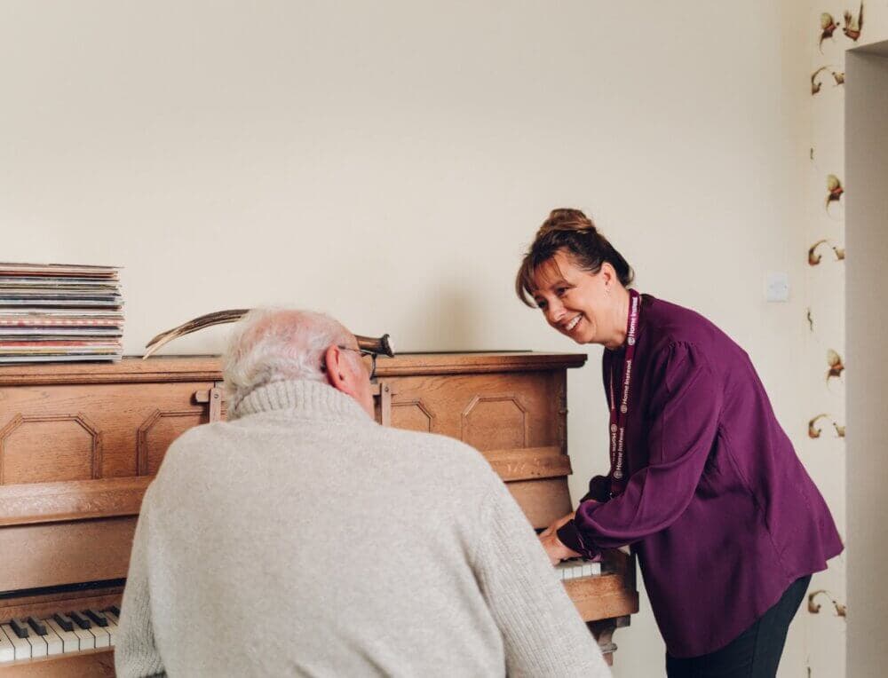 A woman smiles as she helps an elderly man play the piano in a cozy room. - Home Instead