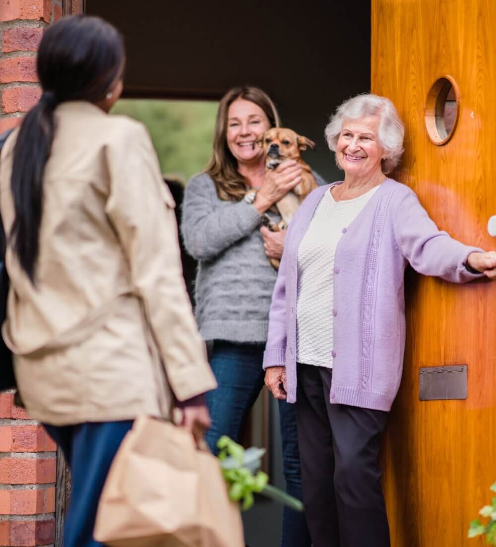 Woman greets two women and a small dog at the door. One woman holds a grocery bag, while another smiles, holding the dog. - Home Instead