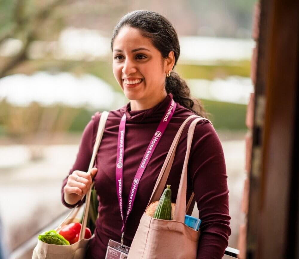 Smiling woman with long dark hair carrying grocery bags and wearing a lanyard, standing outdoors. - Home Instead