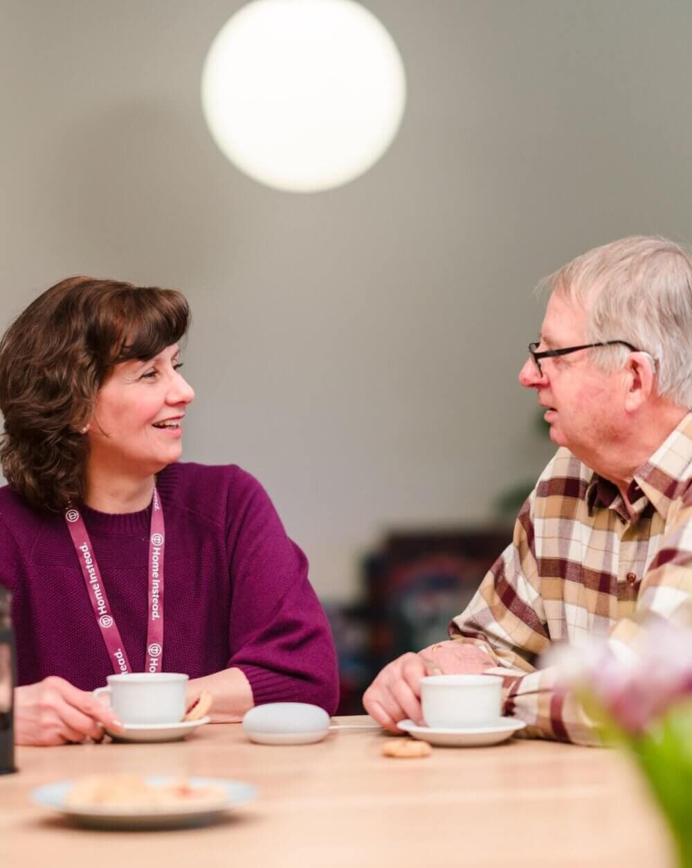 Two people sitting at a table, smiling, sharing a conversation over coffee. - Home Instead