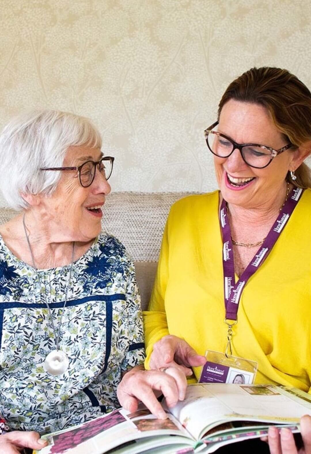 An elderly woman and a caregiver with an ID badge share a joyful moment while looking at a book together. - Home Instead