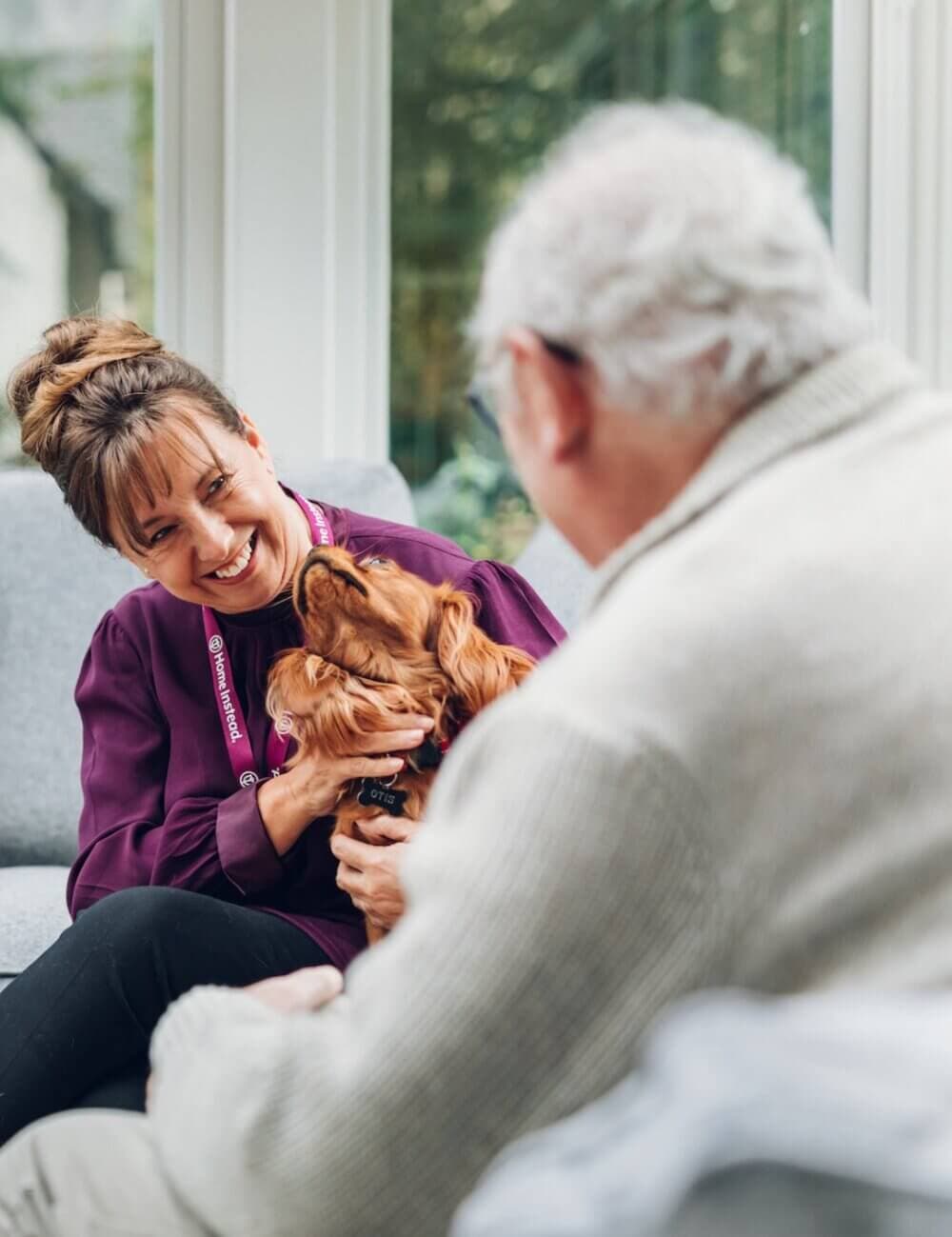 A woman smiles at a man while holding a small dog, both seated in a cozy, well-lit room. - Home Instead